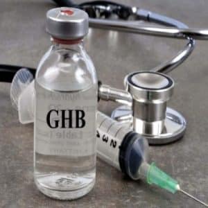 A clear glass vial labeled "GHB" containing a colorless liquid, positioned next to a syringe with a green needle and a stethoscope on a gray surface.