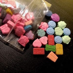 A clear plastic bag filled with various colorful ecstasy tablets, including heart shapes and other designs, scattered on a dark surface.