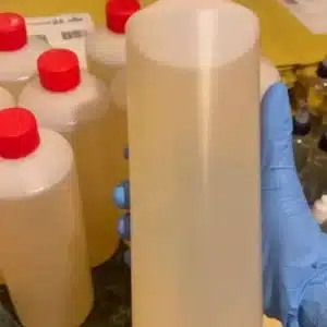 A gloved hand holds a clear plastic bottle filled with a light yellow liquid, surrounded by several similar bottles with red caps on a countertop.