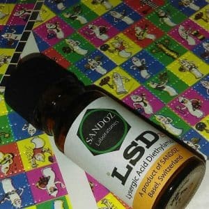 A small brown bottle labeled "LSD" with the text "Lysergic Acid Diethylamide" and "A product of SANDOZ, Basel, Switzerland," placed on a colorful background featuring cartoon characters and patterns.
